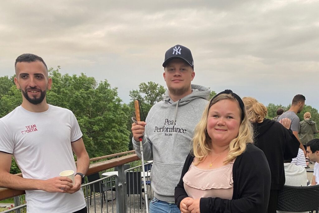 Tre personer står utomhus. En håller en kaffemugg och en håller i en vit käpp.