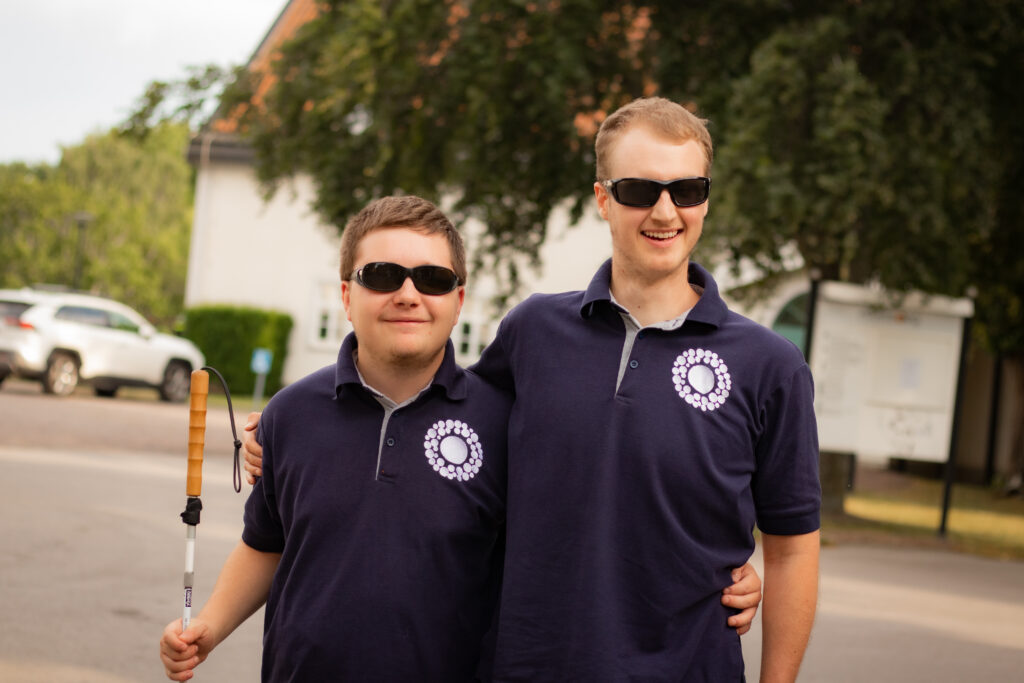 Två leende killar står och håller armen om varandra. Båda bär t-shirt med Unga med synnedsättnings logotyp.
