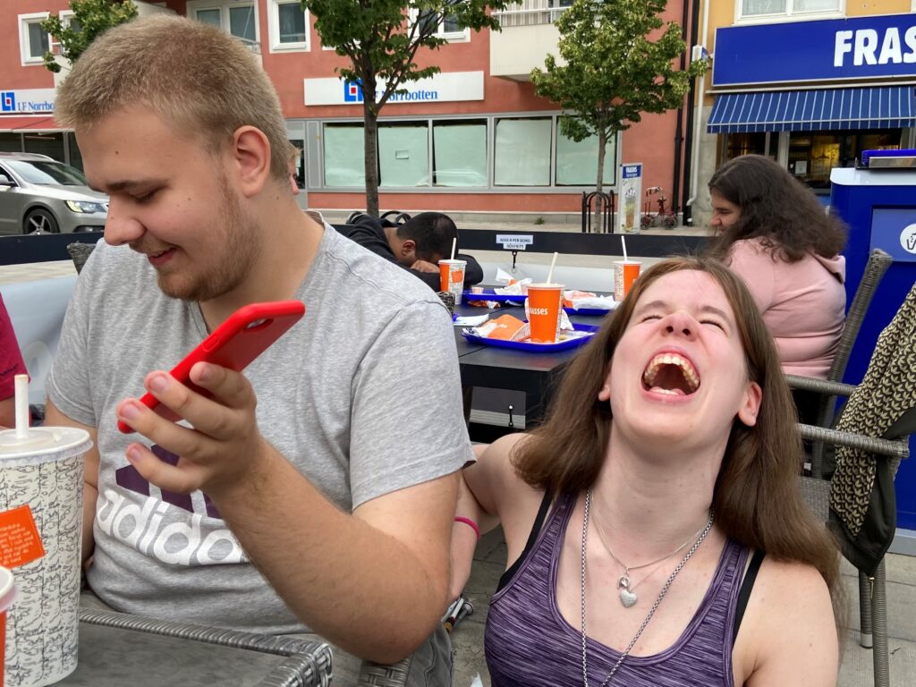 Två skrattande ungdomar på en utomhusservering. En av dem håller i en mobil.