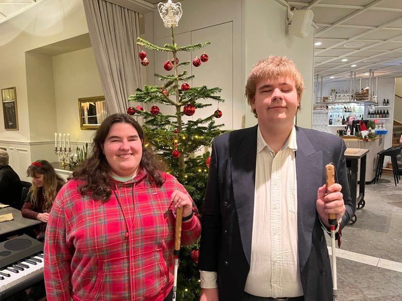 Två ungdomar står i en festlokal med en julgran bakom sig. Båda håller i en vit käpp.